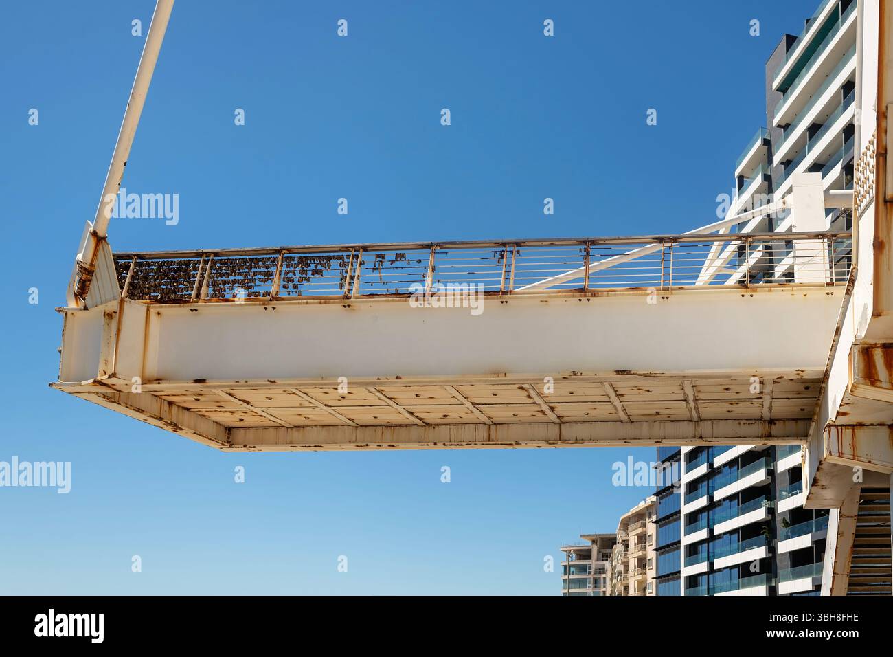 Vista dal basso di un ponte pedonale arrugginito con lucchetti d'amore sulla ringhiera, edifici moderni sullo sfondo e un cielo azzurro. Foto Stock