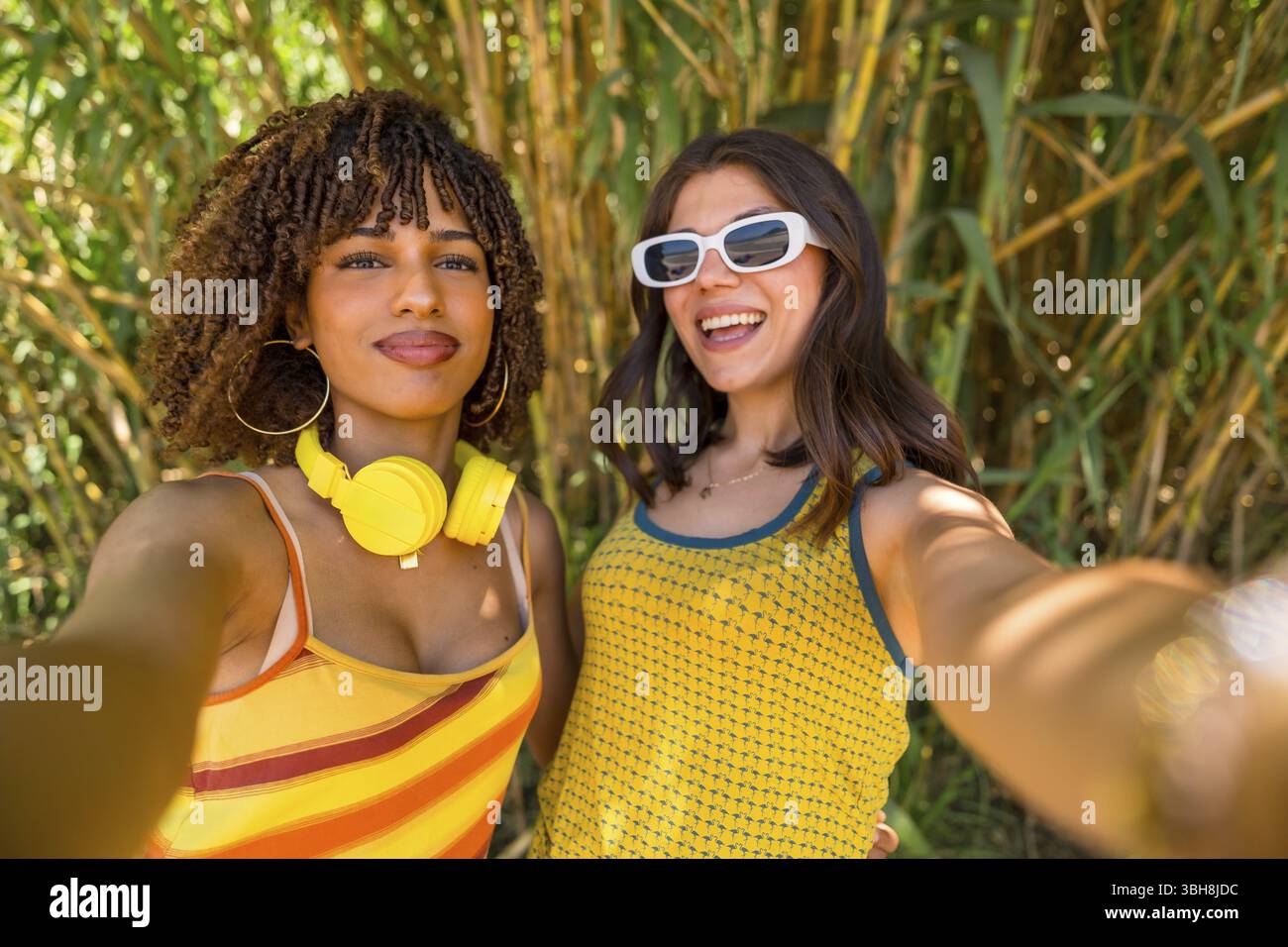 Due giovani donne allegre con abiti colorati e cuffie che scattano un selfie di fronte a una foresta di bambù Foto Stock