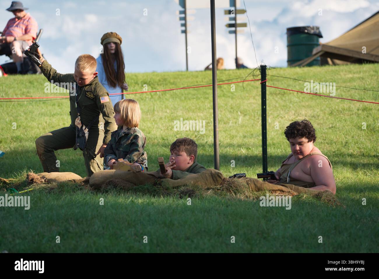 Quattro bambini, uno in tuta militare, giocano con le pistole giocattolo in un campo erboso vicino ai sacchi di sabbia. Una donna e un uomo sono visibili sullo sfondo. Foto Stock