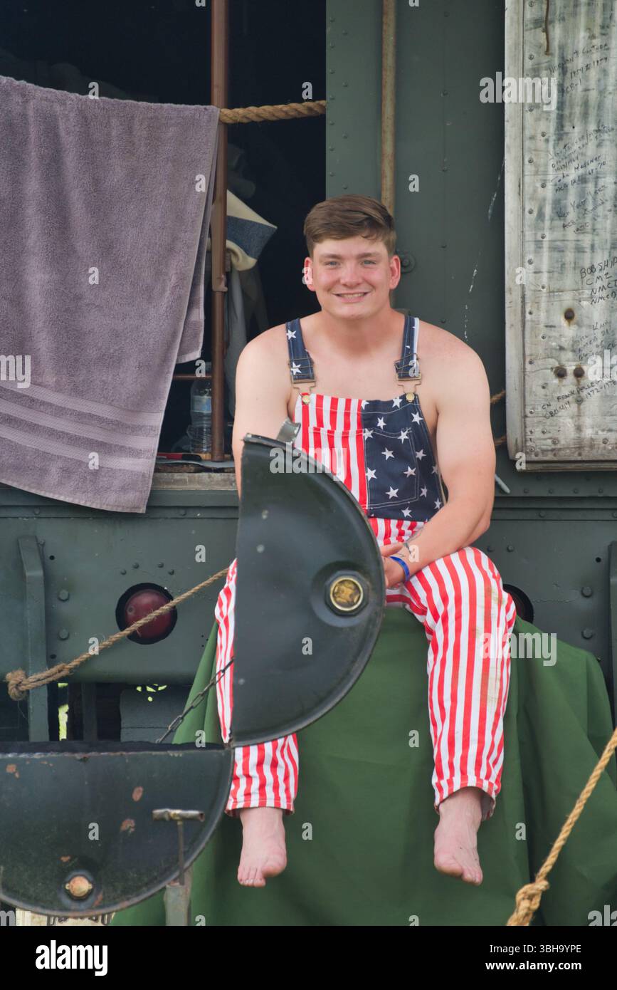 Un giovane uomo dalla pelle leggera siede a piedi nudi su un panno verde, indossando tute a righe rosse e bianche. E' posizionato di fronte a un vefico verde militare Foto Stock