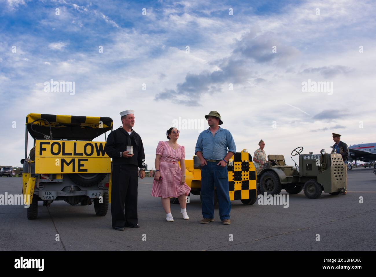Quattro persone in abbigliamento d'epoca si trovano vicino a veicoli militari d'epoca in un campo d'aviazione. Un veicolo Follow me è in primo piano. Il cielo è in parte clou Foto Stock