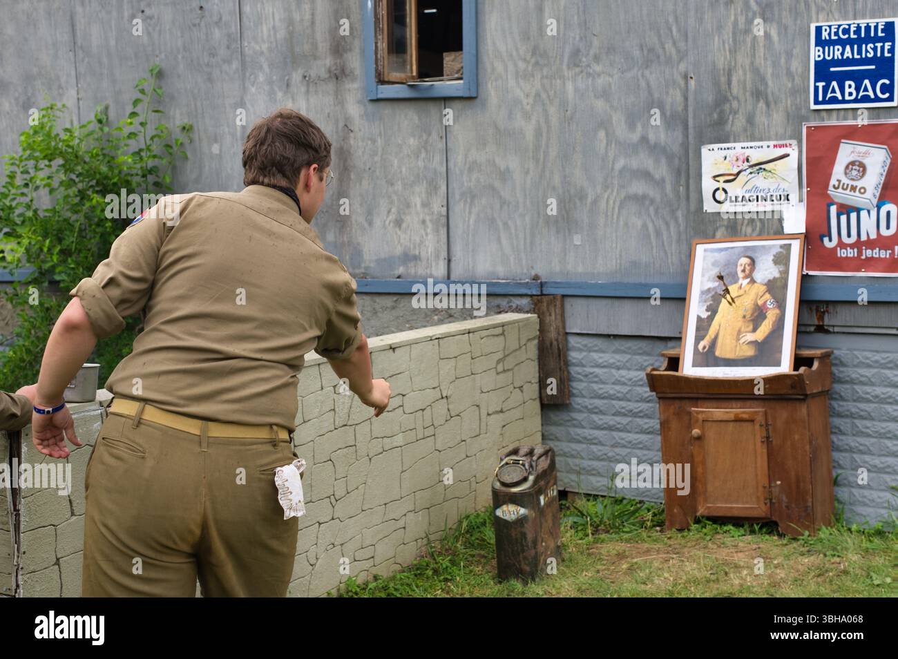 Una persona con una camicia e pantaloni in stile militare abbronzata si trova di fronte a un basso muro di pietra, con pubblicità vintage e un ritratto incorniciato di Adolf Hitler Foto Stock
