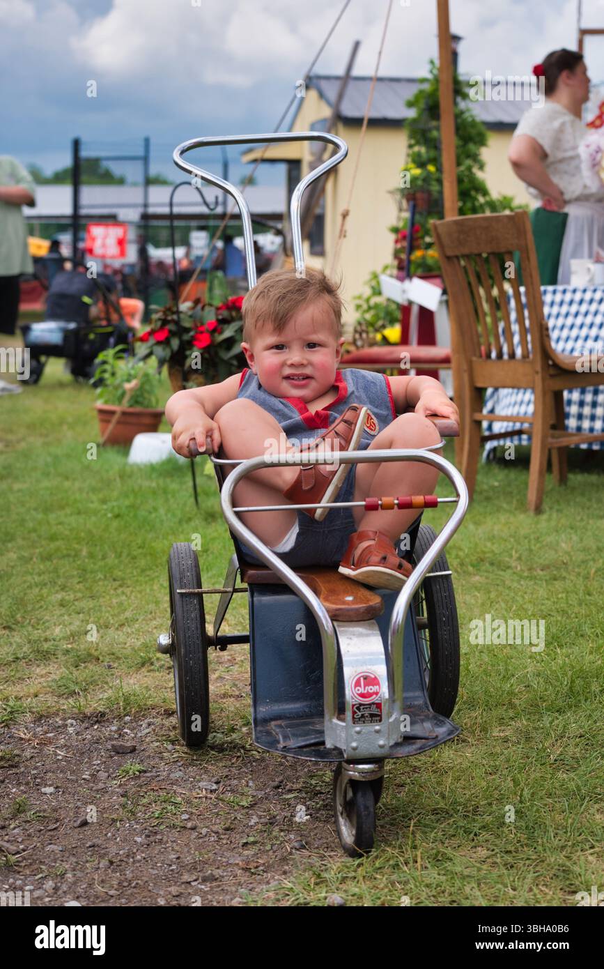 Un bambino siede su un carro in metallo vintage con dettagli in legno. Il carro è grigio scuro/blu e ha una maniglia cromata. Il bambino indossa sandali AN Foto Stock