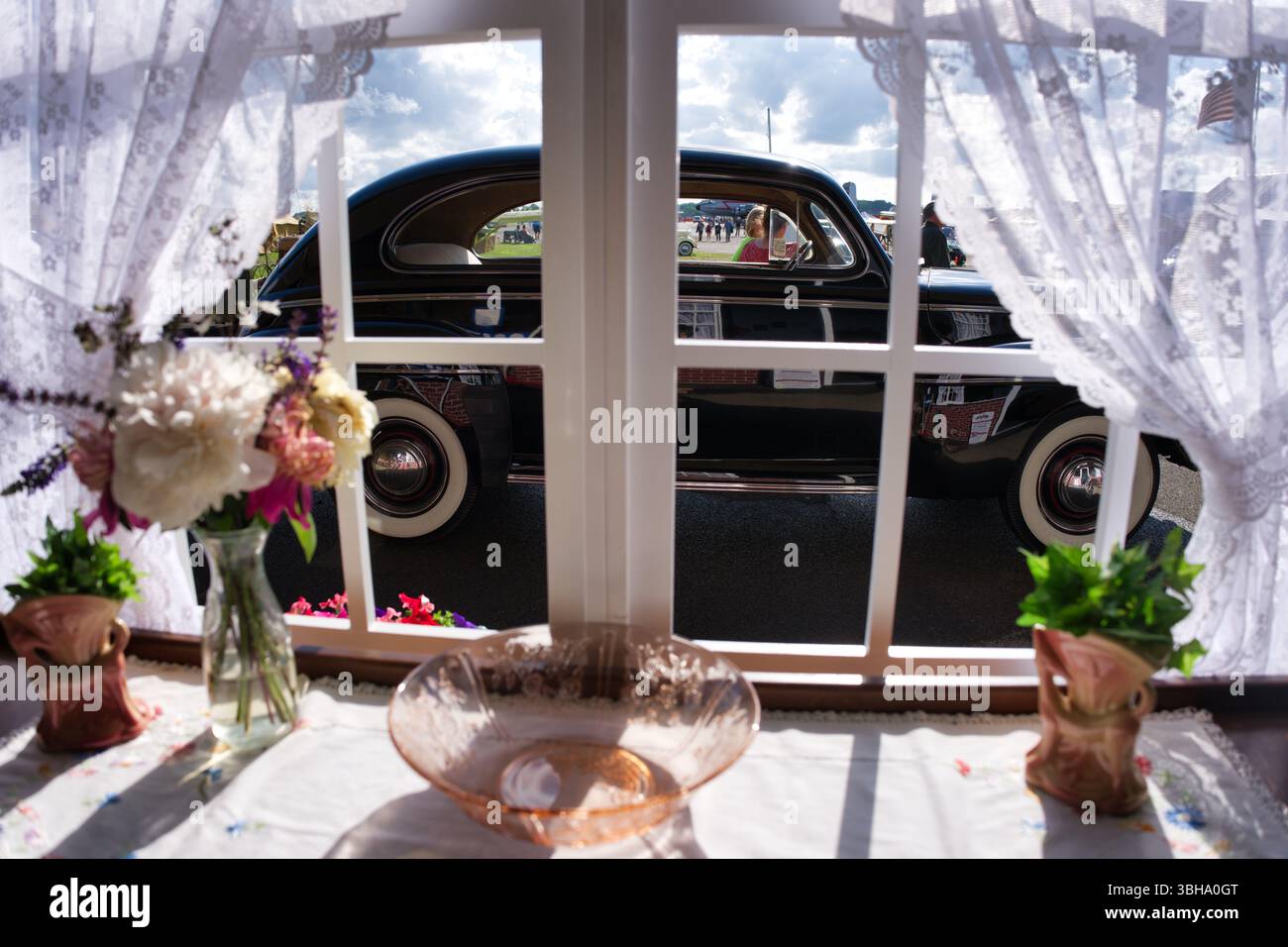 Un'auto nera d'epoca viene vista attraverso una finestra con tende in pizzo. Fiori in vasi e una ciotola di vetro si trovano su un tavolo davanti alla finestra. Luce solare Foto Stock