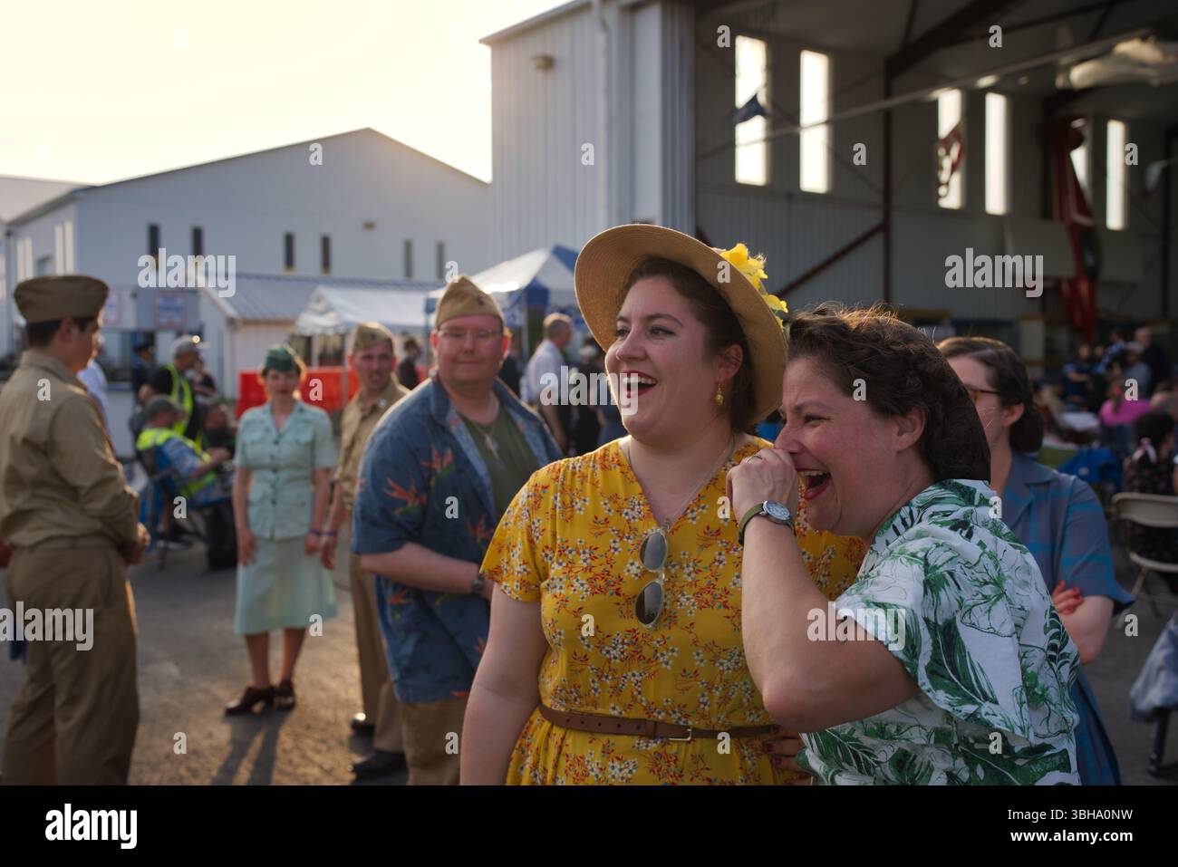 Due donne con abiti vintage anni '1940 ridono insieme all'aperto a un raduno di persone, alcune in stile militare. Un edificio simile a un hangar sono io Foto Stock