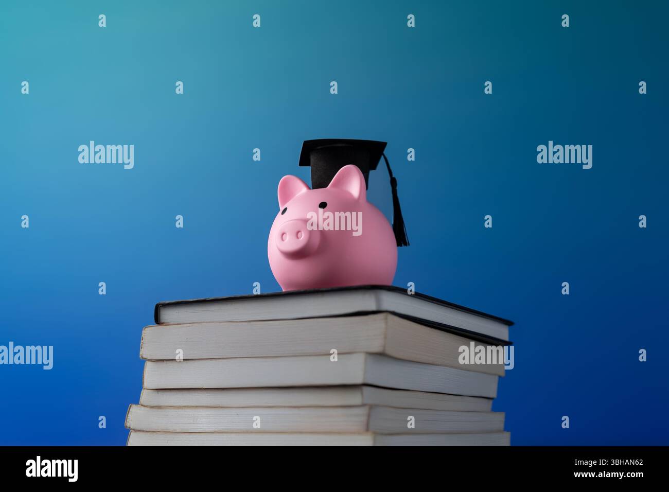 Un salvadanaio rosa che indossa un berretto nero graduato sopra una pila di libri su uno sfondo blu. Risparmiare per l'istruzione, la pianificazione finanziaria Foto Stock