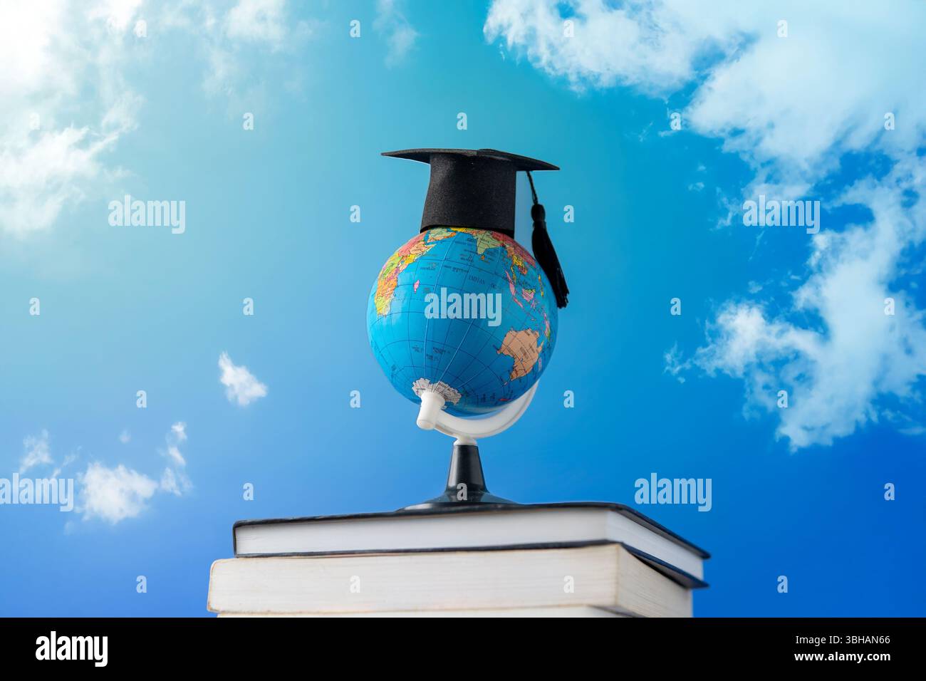 Un globo che indossa un cappello graduato sopra una pila di libri contro un cielo blu vibrante. Istruzione globale, apprendimento internazionale, conoscenza Foto Stock