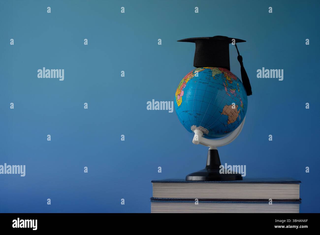 Un globo che indossa un berretto nero graduato su una pila di libri su uno sfondo blu. Copia spazio per il testo. Istruzione globale, accademica Foto Stock