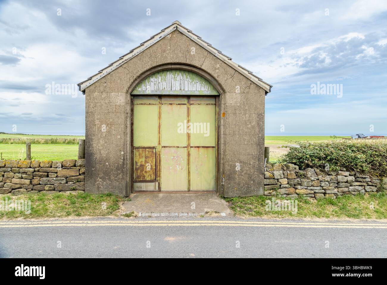 Un edificio solitario a lato della strada incorniciato da un muro di pietra in un'ampia campagna aperta, che cattura l'isolamento tranquillo e la semplicità della vita rurale. Foto Stock