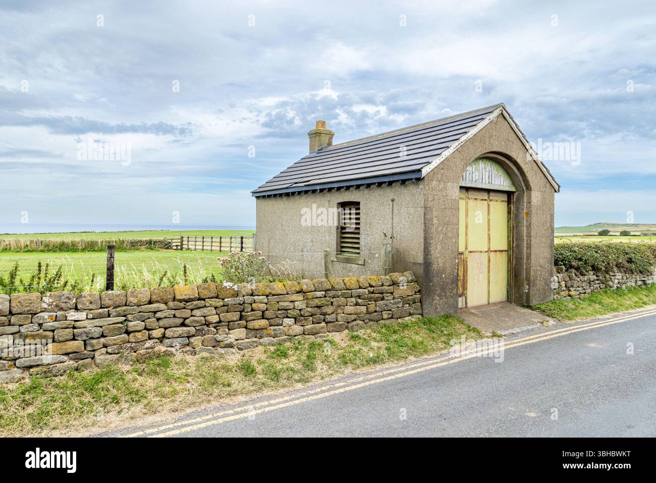 Un edificio solitario a lato della strada incorniciato da un muro di pietra in un'ampia campagna aperta, che cattura l'isolamento tranquillo e la semplicità della vita rurale. Foto Stock