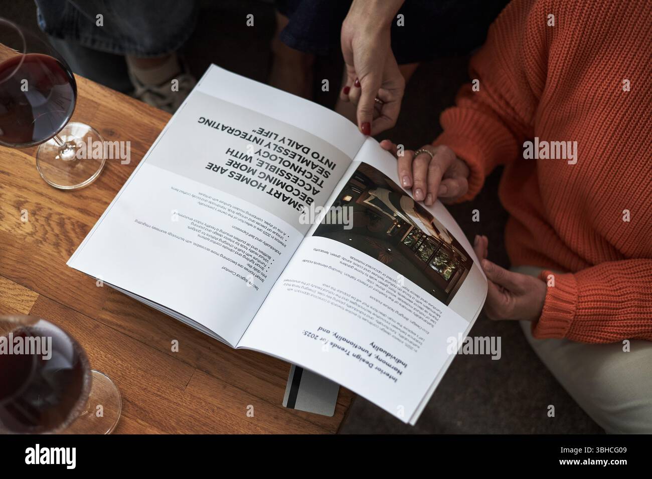 Due giovani donne caucasiche sedute insieme a leggere una rivista aperta su un tavolo di legno, mani visibili che tengono pagine, bicchieri di vino rosso nelle vicinanze Foto Stock