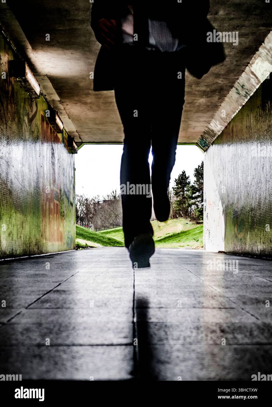 Una persona, presumibilmente un uomo d'affari basato sul suo abbigliamento, sta attraversando un sottopassaggio. Il sottopassaggio è graffeggiato e si apre su un'area erbosa con t Foto Stock
