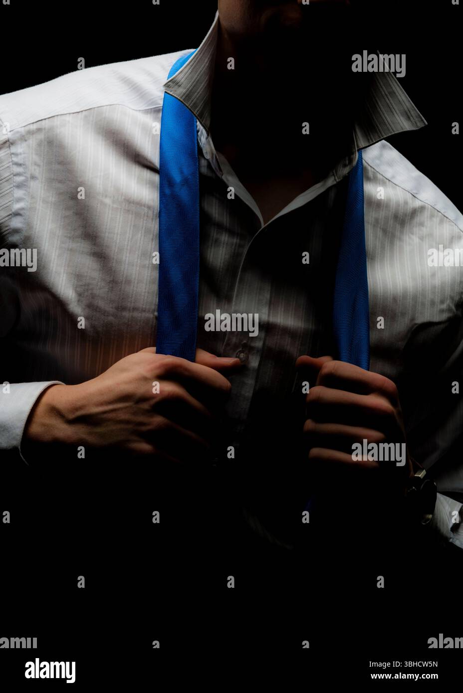 Un uomo con una camicia bianca in pinstriping allenta la cravatta blu a motivo chevron. Il suo volto è oscurato dall'ombra, e lo sfondo è nero. Foto Stock