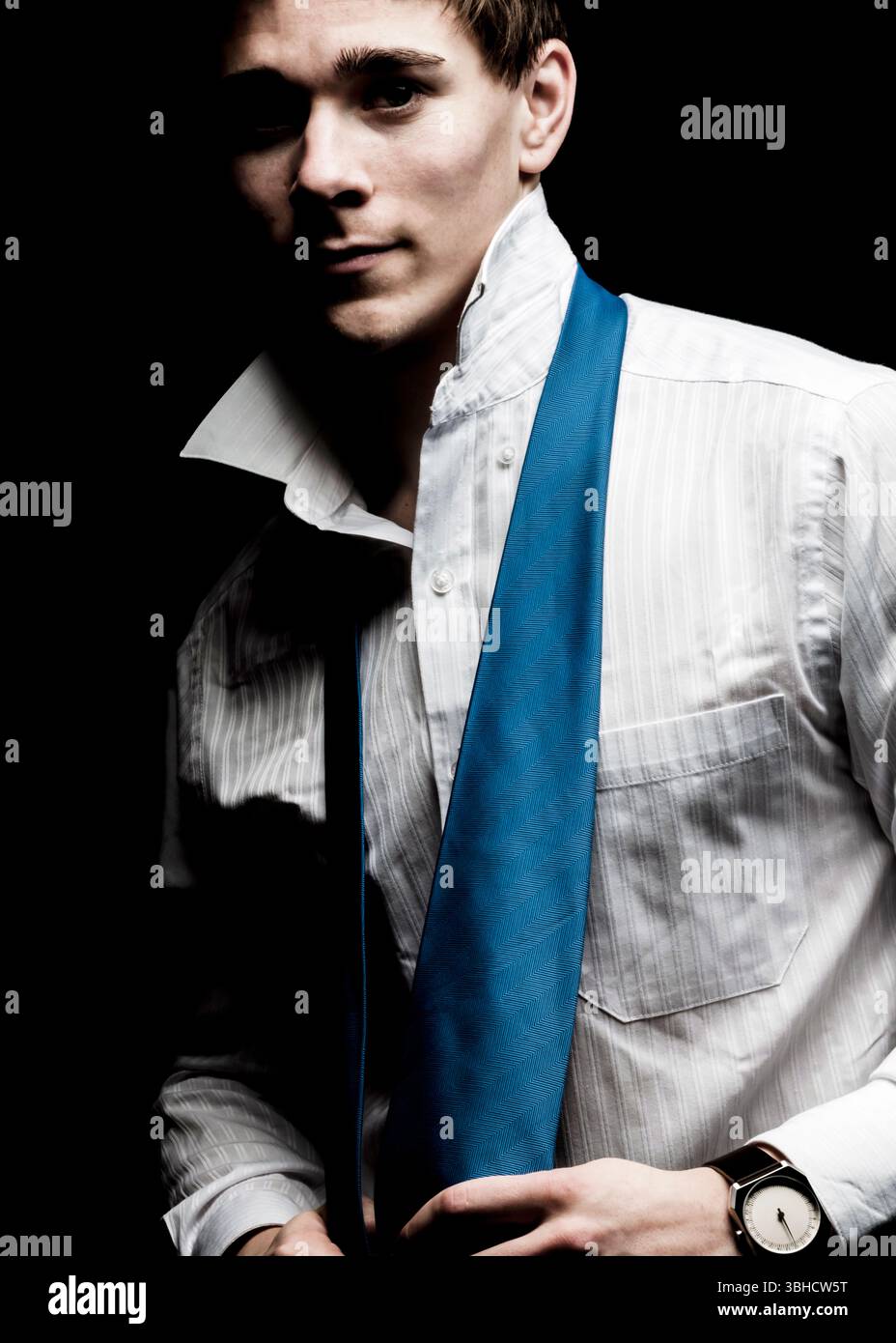 Un uomo con una camicia bianca in tessuto con colletto aperto adatta la sua cravatta blu su uno sfondo nero. Indossa un orologio da polso con un fratello Foto Stock