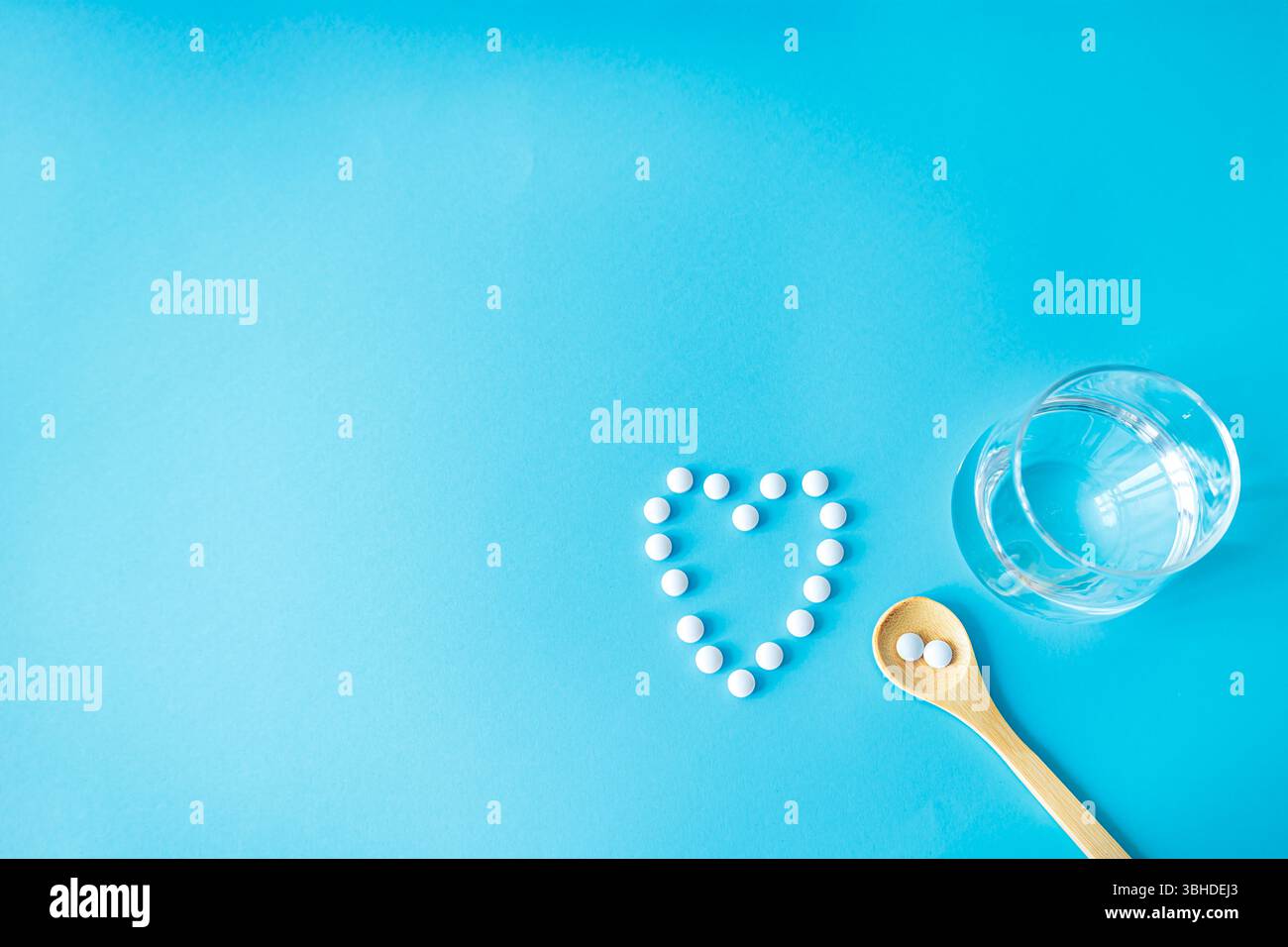 Tavolette bianche a forma di cuore, bicchiere d'acqua e cucchiaio di legno. Appartamento giaceva su sfondo blu simboleggiando l'amore, la cura, la medicina e la routine di salute quotidiana Foto Stock