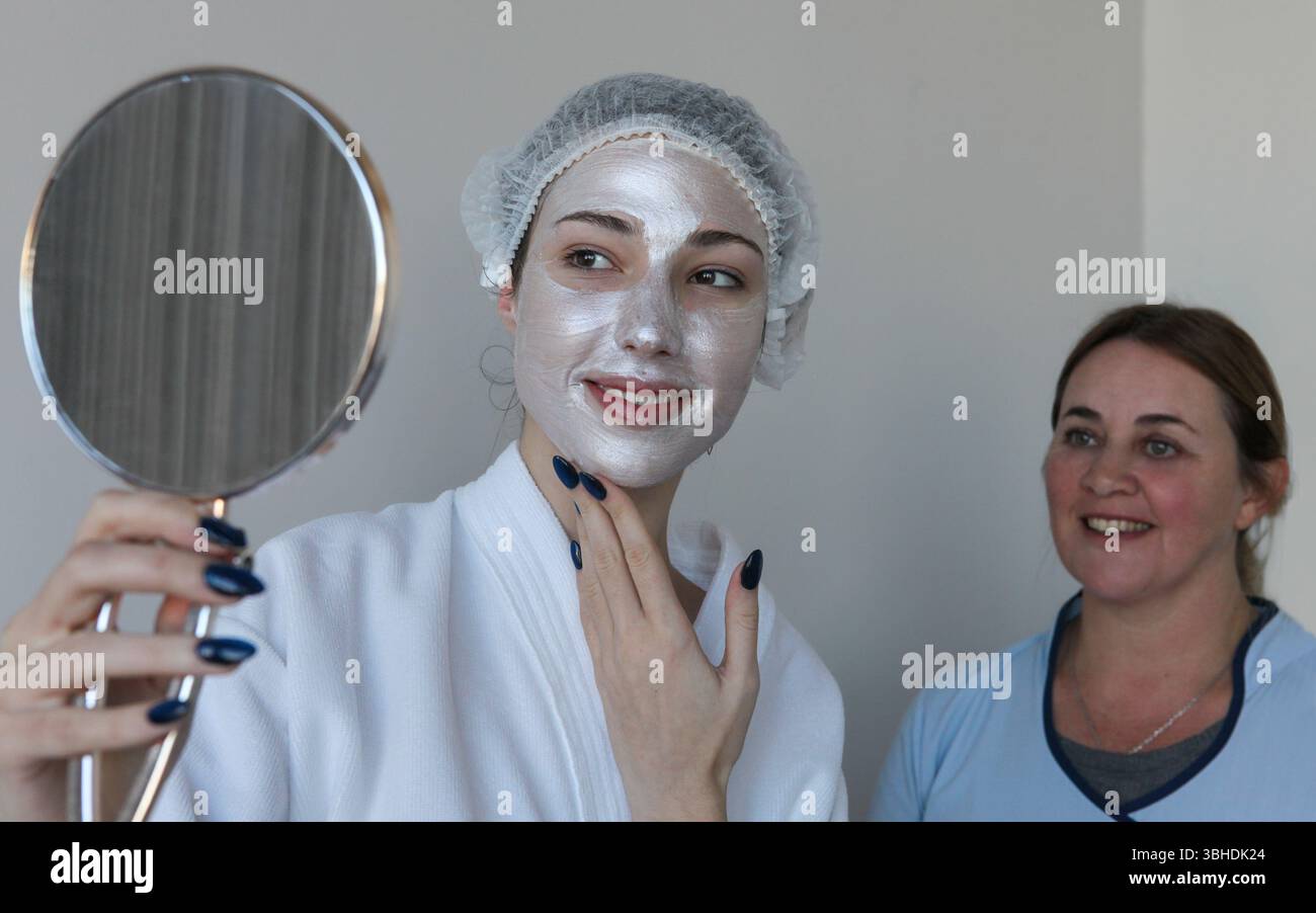 Giovane donna con maschera facciale sorridente mentre guarda uno specchio per le mani al salone spa, estetista in piedi dietro di lei sorridente felicemente, mostrando una gioia Foto Stock