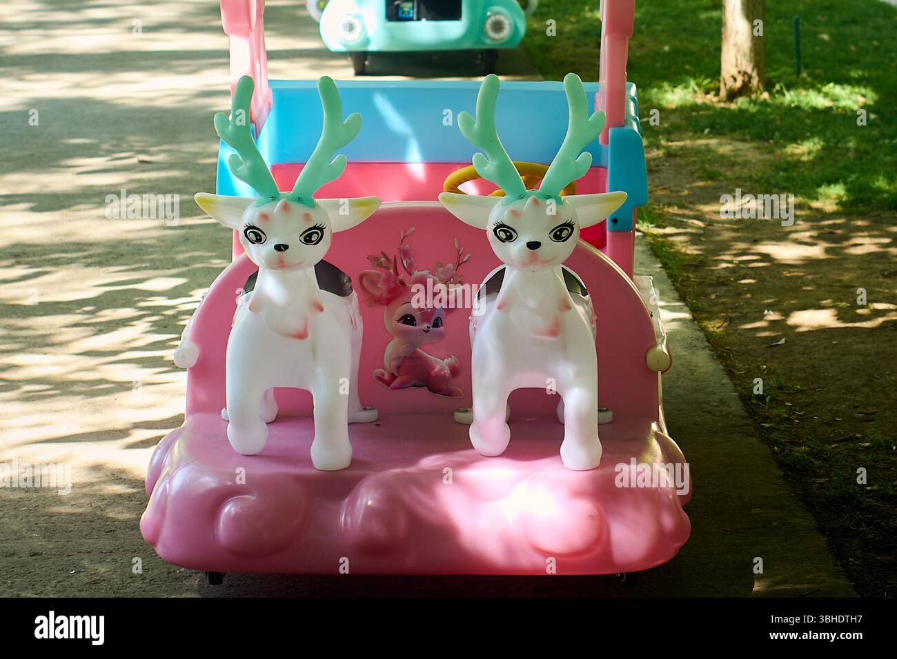 Due cervi di plastica con corna luminose siedono su un carretto rosa e blu, parte di un'attrazione per bambini situata su un sentiero soleggiato nel parco Foto Stock