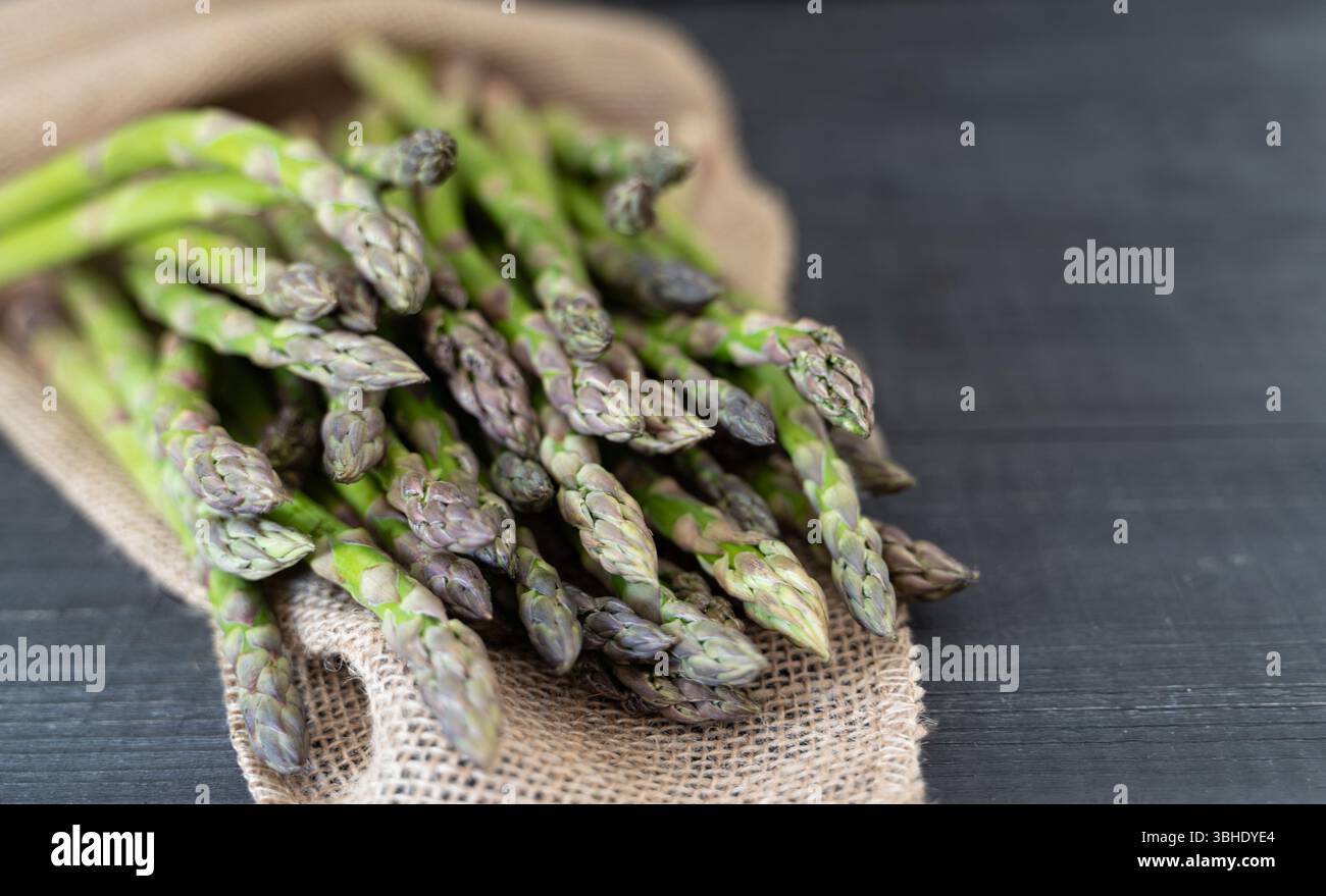 Un fascio di asparagi verdi su una superficie di legno scuro con un sacco grezzo che evoca un'atmosfera fresca di fattoria. Foto Stock