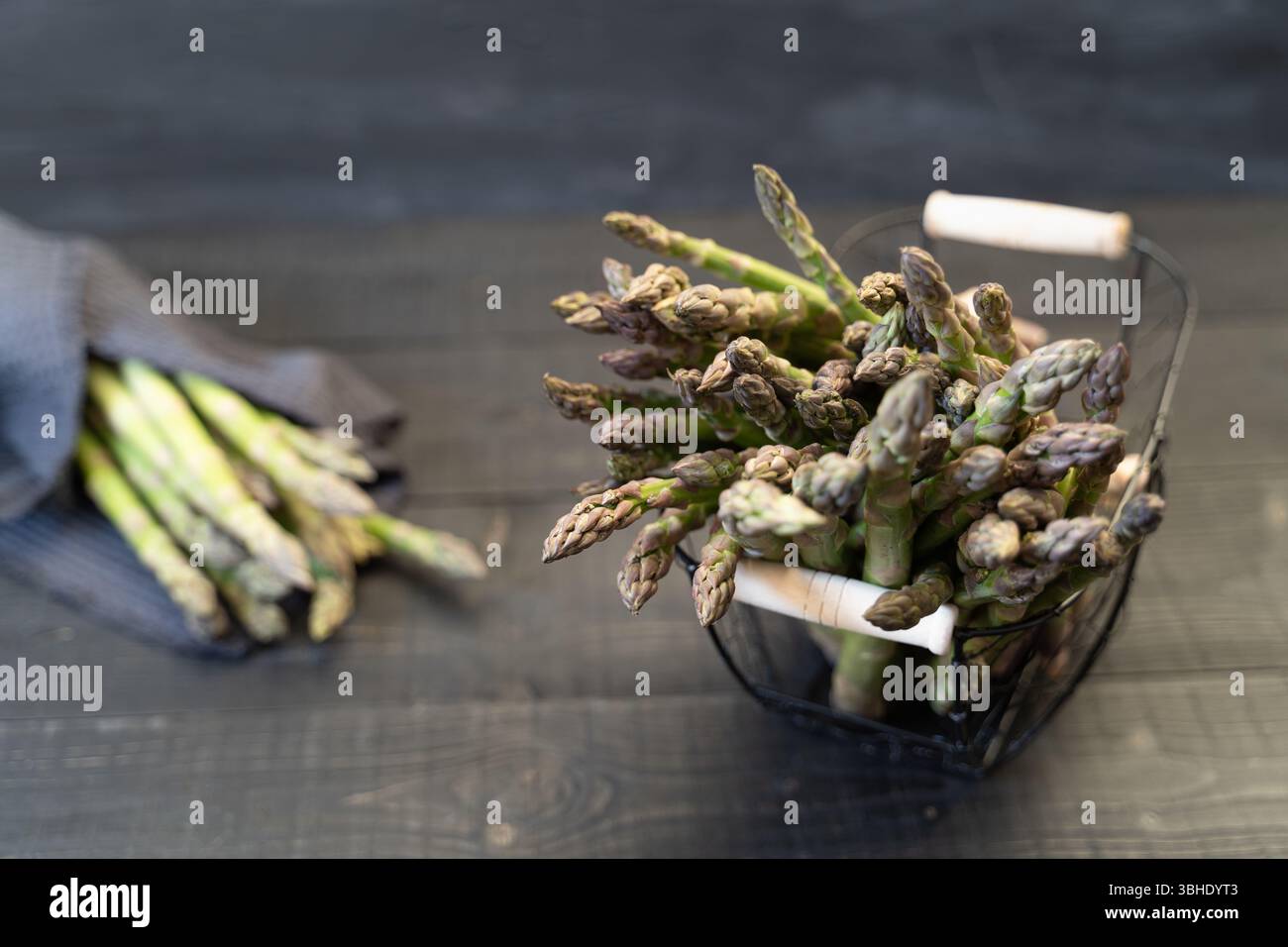Vista dall'alto di asparagi verdi freschi in cestello di filo metallico con un fascio avvolto in un panno da cucina su una superficie in legno nero Foto Stock
