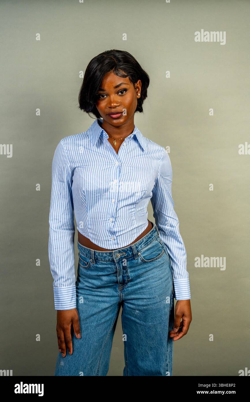 Ritratto in studio di una giovane donna nera con i capelli corti, che indossa una camicia e un jeans a righe blu alla moda, trasudando sicurezza e stile Foto Stock