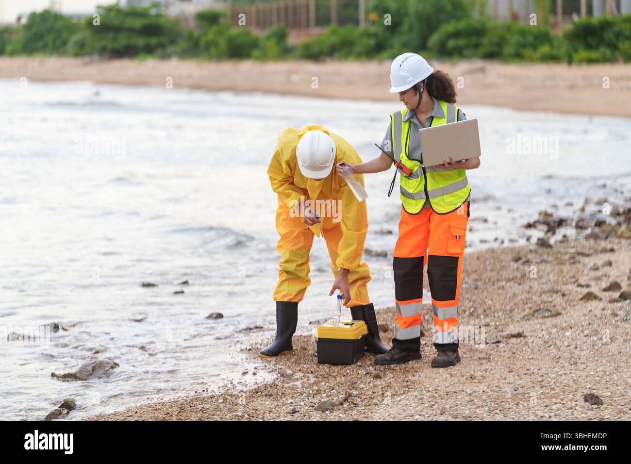 Due professionisti in attrezzature di sicurezza analizzano i campioni d'acqua lungo la spiaggia, garantendo la sicurezza ambientale in una giornata di sole in acqua. Lavorano collaborano Foto Stock