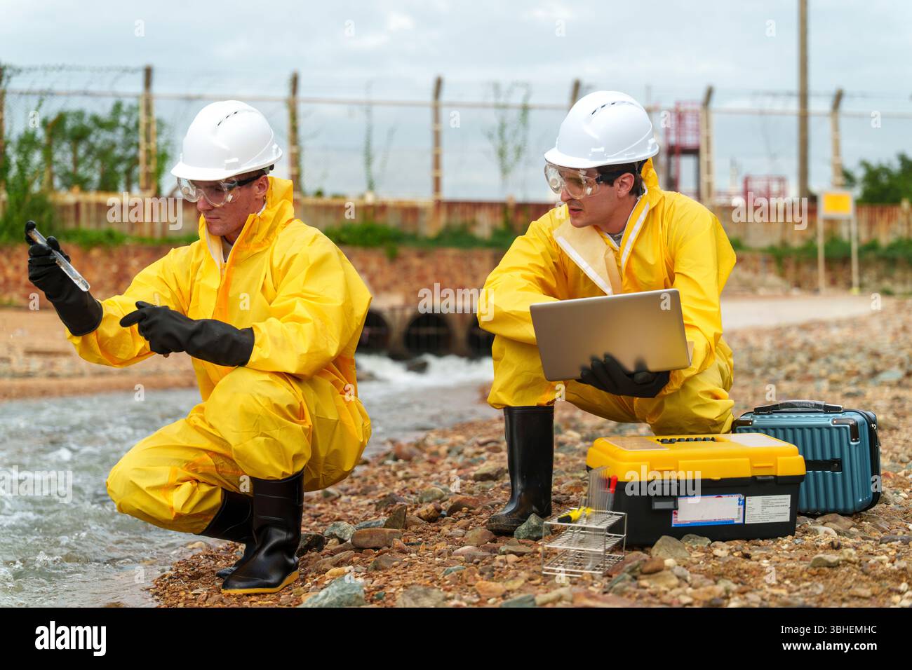 Due scienziati in tute protettive gialle e caschi raccolgono campioni d'acqua da una sponda fluviale. Lavorano con attenzione, utilizzando attrezzature per garantire la massima precisione Foto Stock