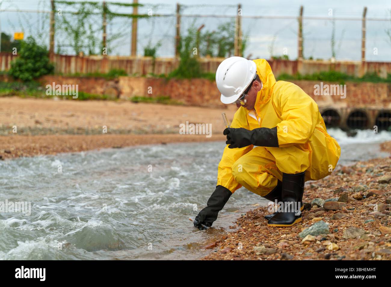 Una persona con tuta da pioggia gialla e attrezzatura di sicurezza accovacciata lungo un fiume, raccogliendo meticolosamente campioni d'acqua per la ricerca ambientale sulla contaminazione i. Foto Stock