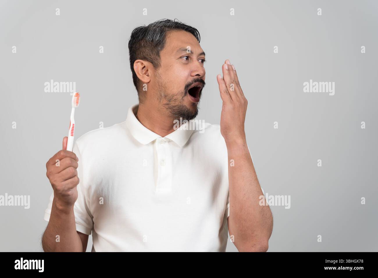 Uomo asiatico di mezza età che controlla il respiro con la mano vicino alla bocca mentre tiene in mano lo spazzolino da denti, concetto di igiene orale, problema di alito cattivo, routine mattutina, Foto Stock