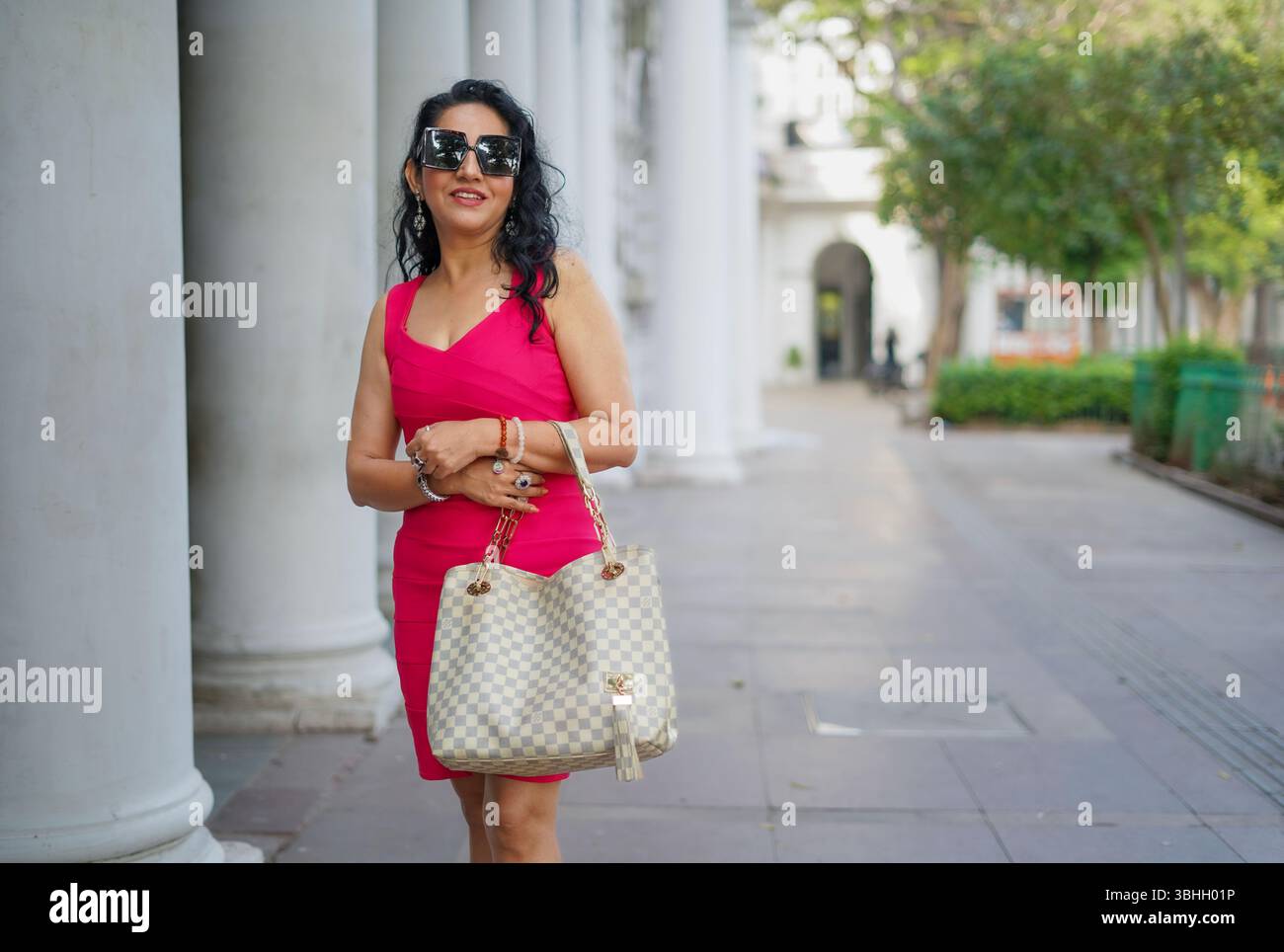 Donna elegante con un abito rosa brillante che porta con sé una borsa di design mentre cammina attraverso una passeggiata in città, la moda e il concetto di stile urbano moderno. Foto Stock