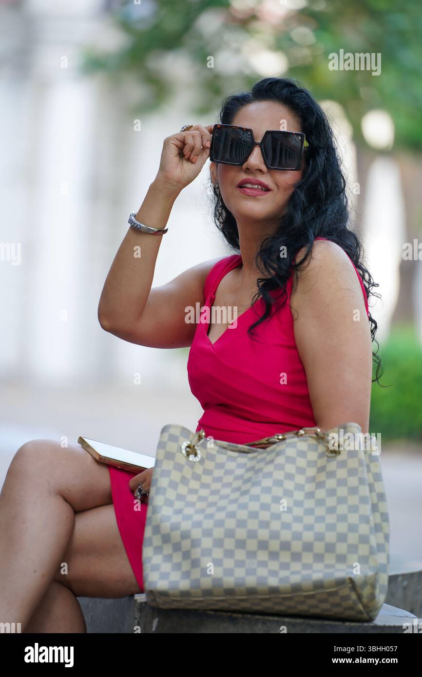 Donna elegante con un abito rosa brillante che porta con sé una borsa di design mentre cammina attraverso una passeggiata in città, la moda e il concetto di stile urbano moderno. Foto Stock