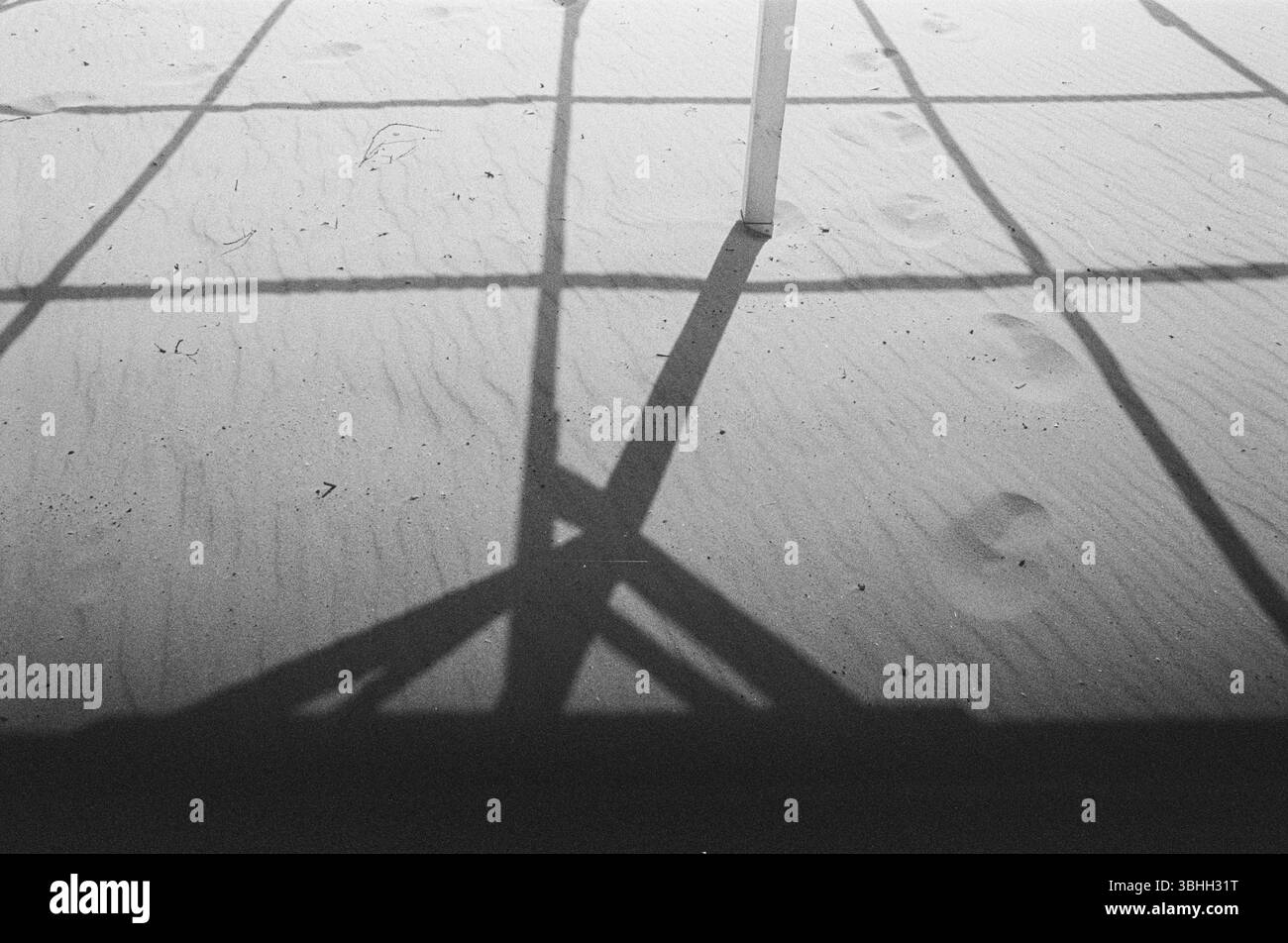 Foto in bianco e nero di ombre e impronte intersecanti sulla sabbia, catturando geometrie minimaliste, solitudine e equilibrio silenzioso in luce naturale Foto Stock