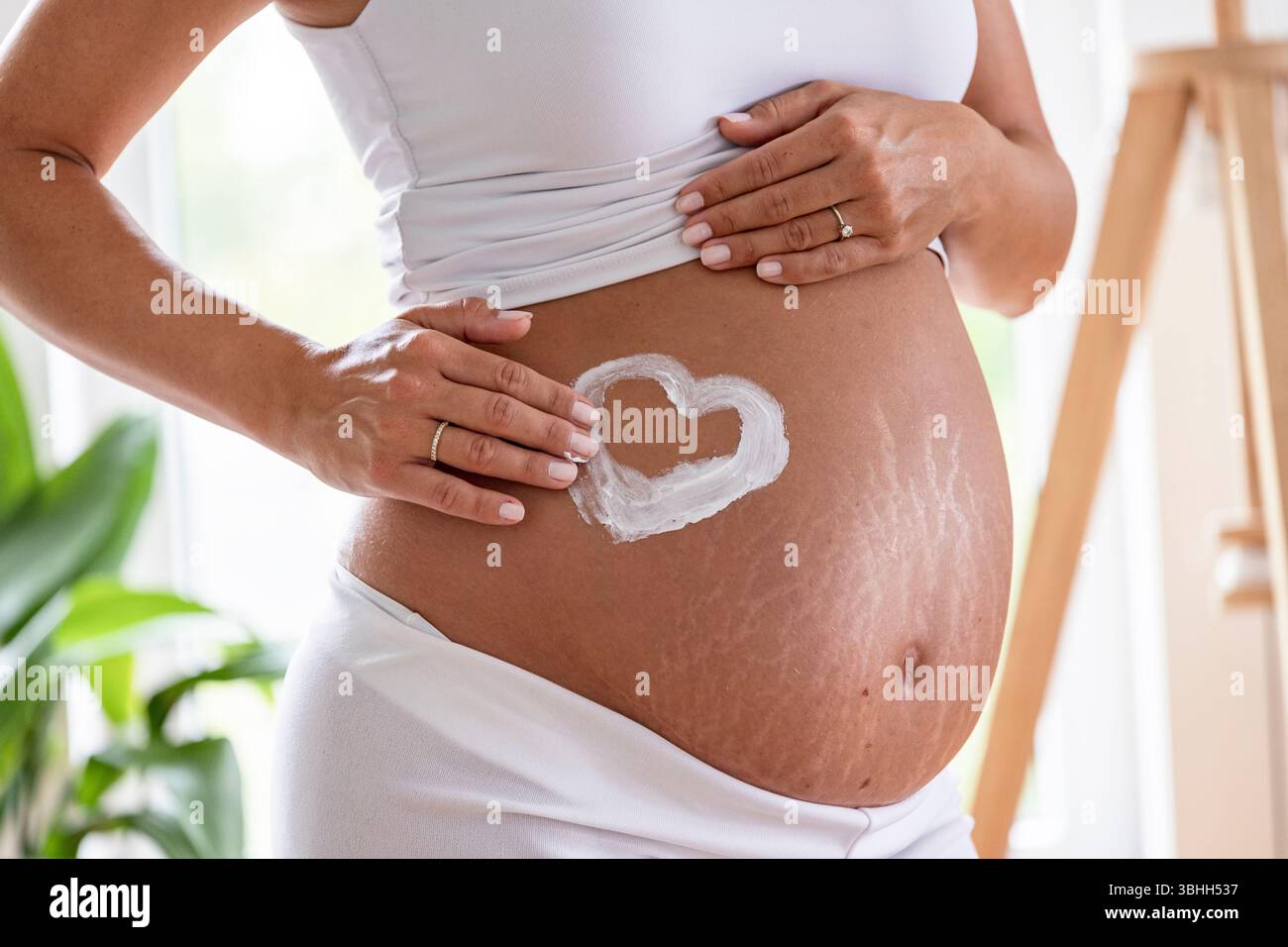 Primo piano di una donna che disegna un cuore con protezione solare sulla pancia incinta, mostrando amore per il suo bambino Foto Stock
