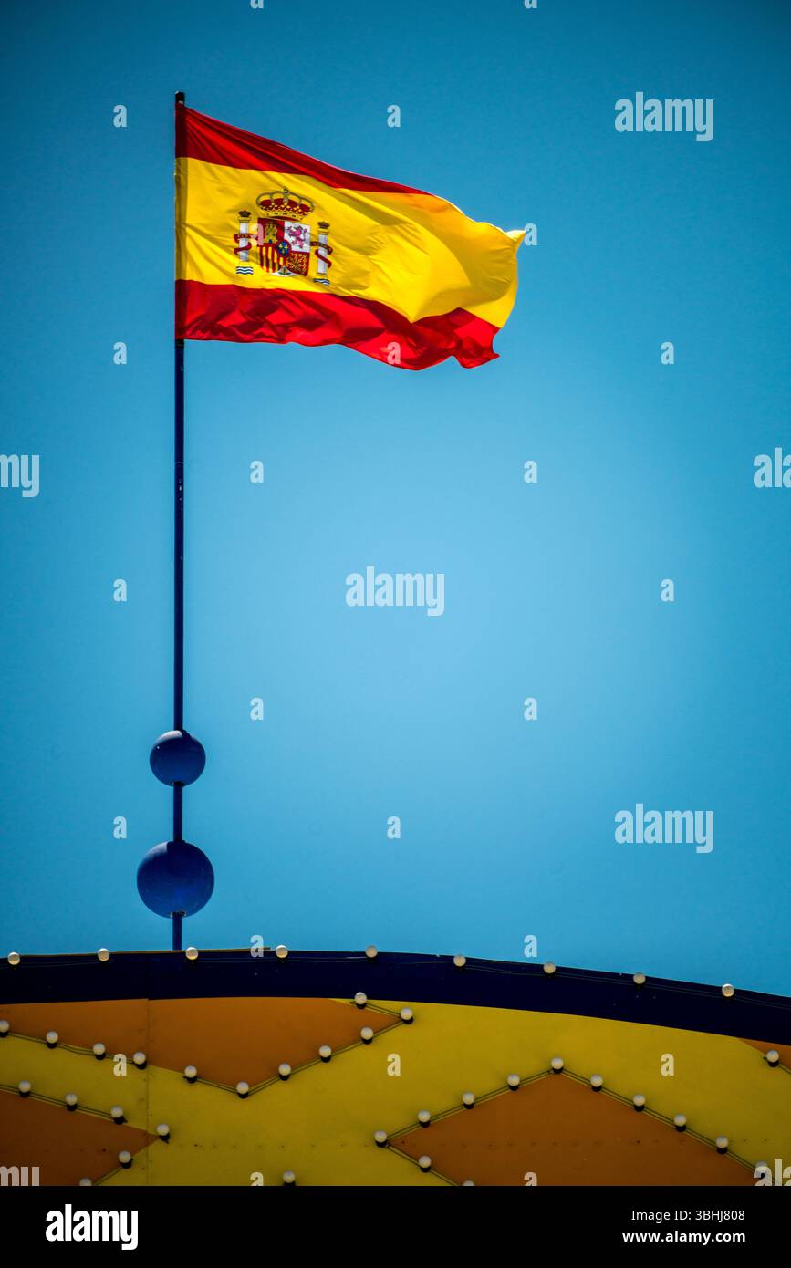 La vibrante bandiera della Spagna ondeggia orgogliosamente sopra il cancello d'ingresso della Fiera di aprile a Siviglia, mostrando l'atmosfera festosa. Foto Stock