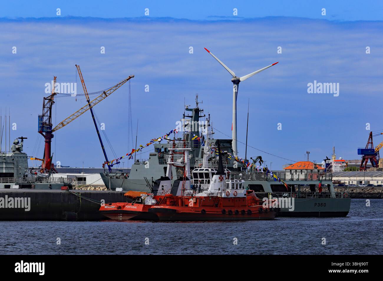 Viana do Castelo, Portogallo - 14 maggio 2025: Rimorchiatori, navi della marina, gru e una turbina eolica collegano il porto industriale sotto un cielo blu Foto Stock
