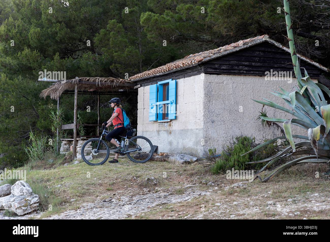 Donna turista in gita in bicicletta, esplorando la penisola di Kamenjak in Istria, Croazia, donna in mountain bike accanto a una casa tradizionale di un pescatore Foto Stock