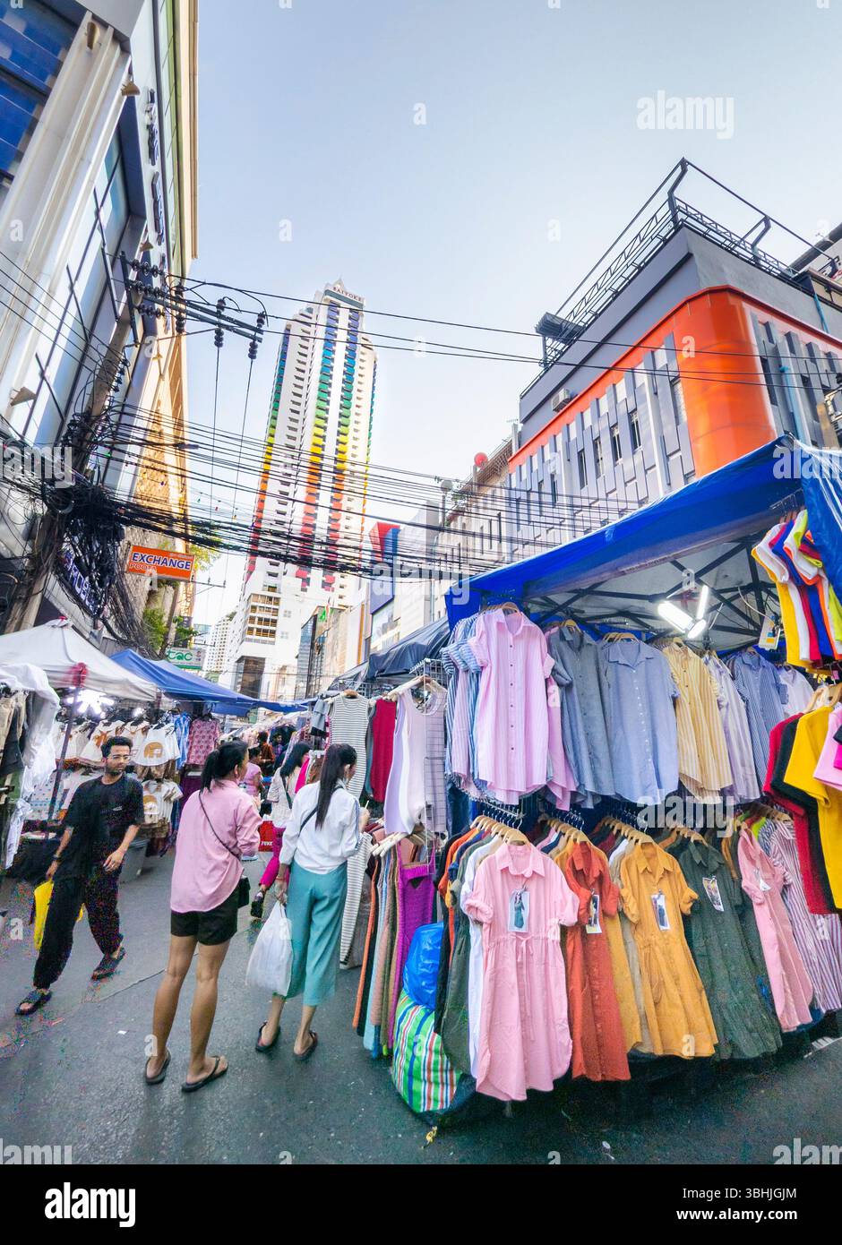 Thailandia - 7 aprile 2024 - persone curiosano tra le scaffalature di abbigliamento, all'aperto a livello della strada, mentre l'iconico edificio condominio, un punto di riferimento della città, si infila in lontananza Foto Stock