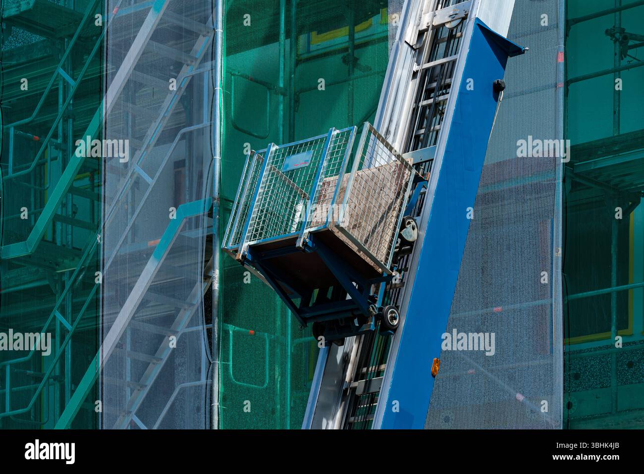 Un cantiere è dotato di un ascensore utilizzato per trasportare materiali da costruzione. L'ascensore è posizionato lungo una struttura alta che mostra il verde saf Foto Stock