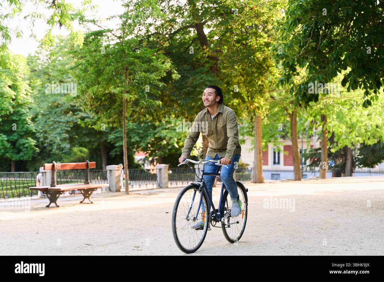 Giovane uomo che va in bicicletta attraverso un vivace parco cittadino verde, prendendo il sole e godendosi una spensierata giornata estiva all'aria aperta Foto Stock