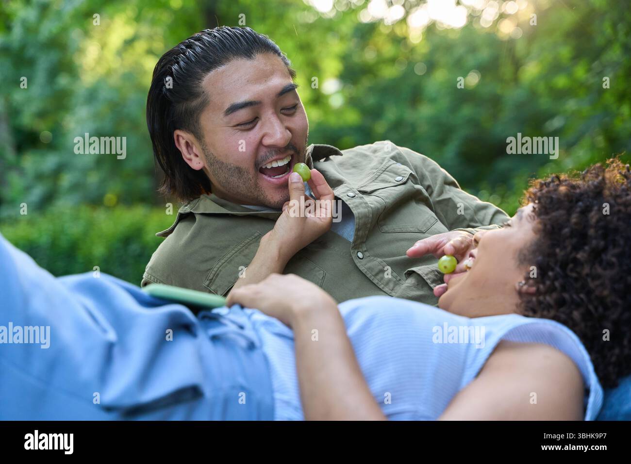 Coppia felice che si diverte durante un romantico picnic in un parco, condividendo l'uva e godendosi il tempo estivo Foto Stock