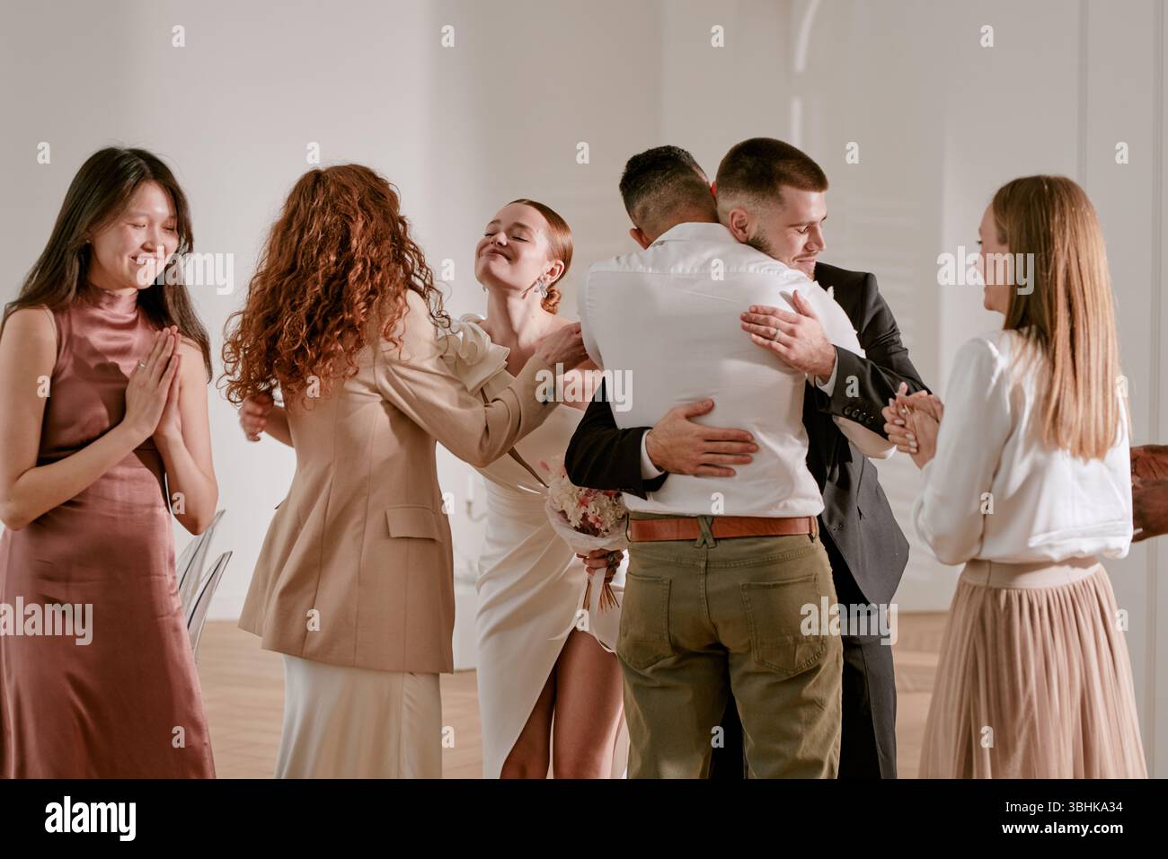 Gruppo di giovani adulti e multietnici che celebrano il matrimonio, sposa che tiene il bouquet sorridente mentre abbraccia lo sposo, amici che si alzano nelle vicinanze applaudono e si congratulano con la coppia Foto Stock