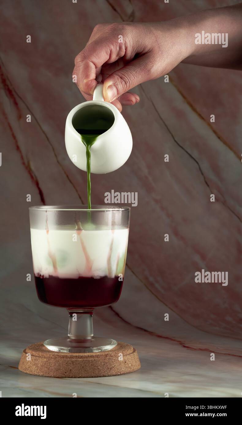 Una mano versa il matcha verde in un bicchiere contenente uno strato rosso di fragola, creando una bevanda visivamente attraente e stratificata Foto Stock