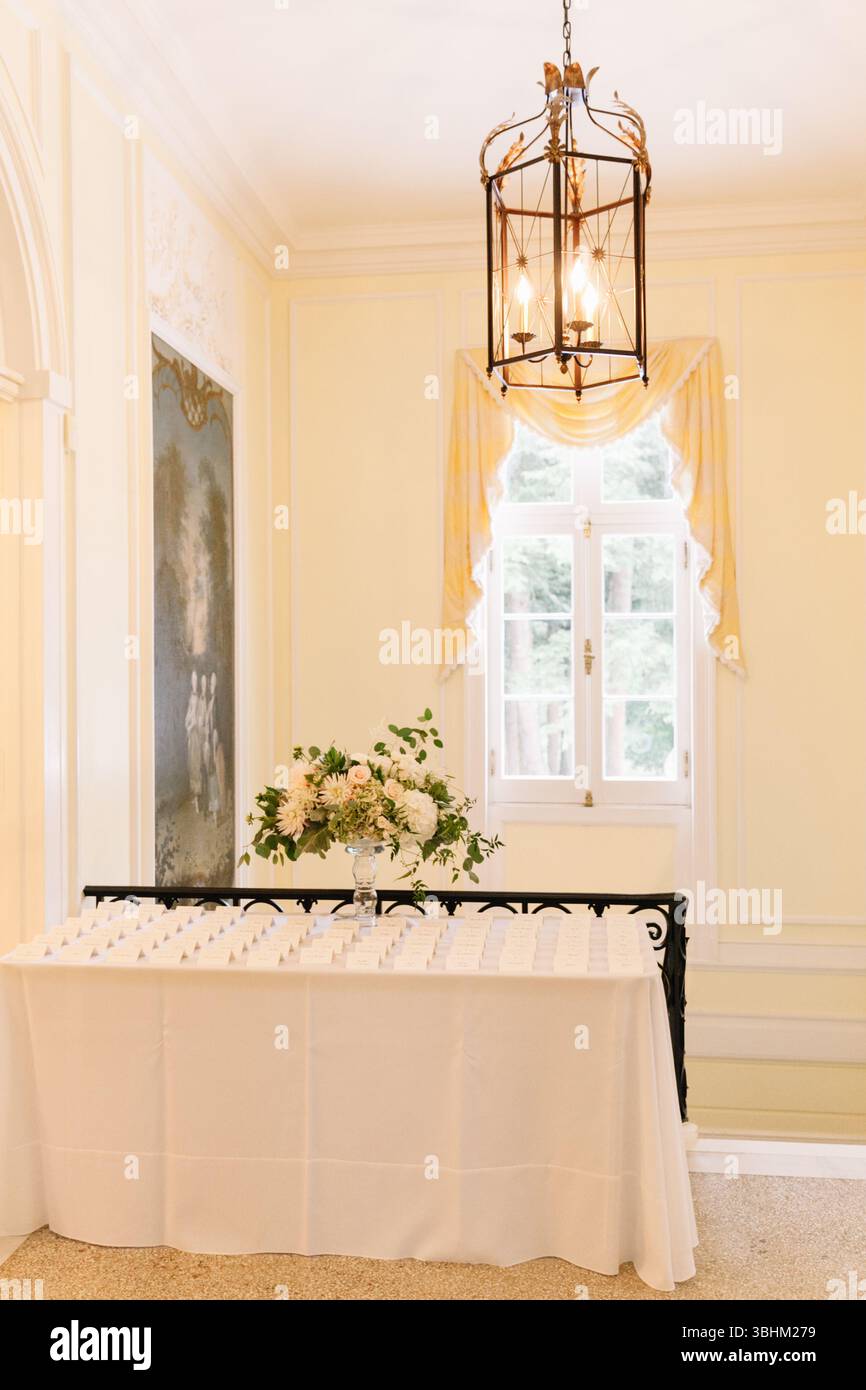 La disposizione floreale decora un tavolo con segnaposto in una sala ben illuminata decorata con mobili eleganti. Foto Stock