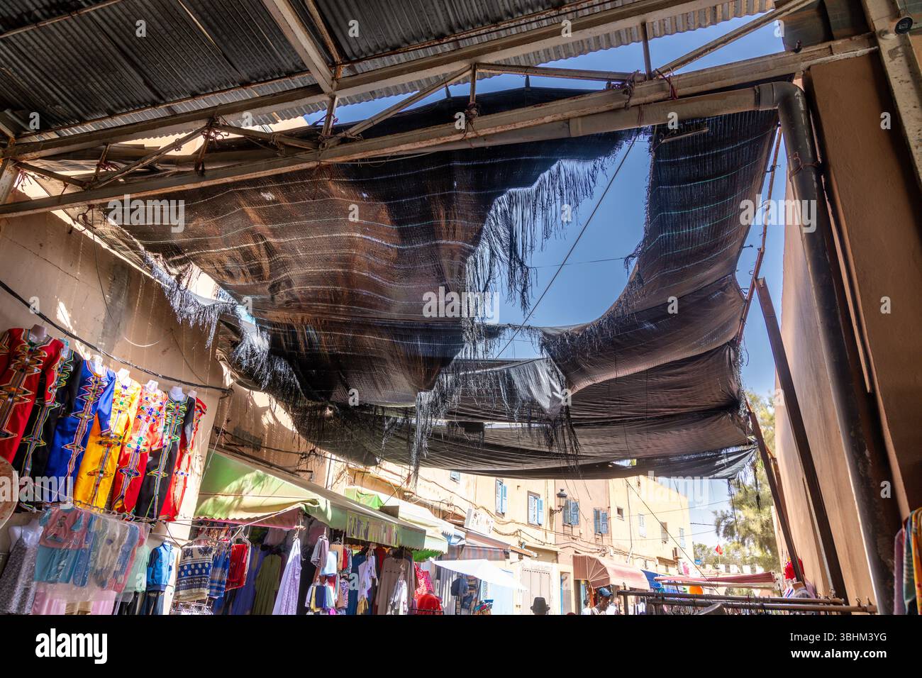 Agadir, Souss-massa - Marocco - 08-03-2024: Area del mercato coperta con una tendina parasole nera danneggiata, vestiti colorati esposti sugli stand e luce solare illu Foto Stock