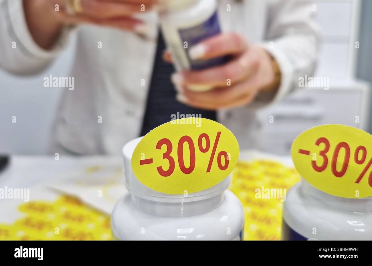 Giovane farmacista donna che indossa uniforme bianca attaccando gli adesivi di sconto agli articoli di medicina da farmacia rifornita o negozio di forniture mediche. Annuncio di marketing Foto Stock