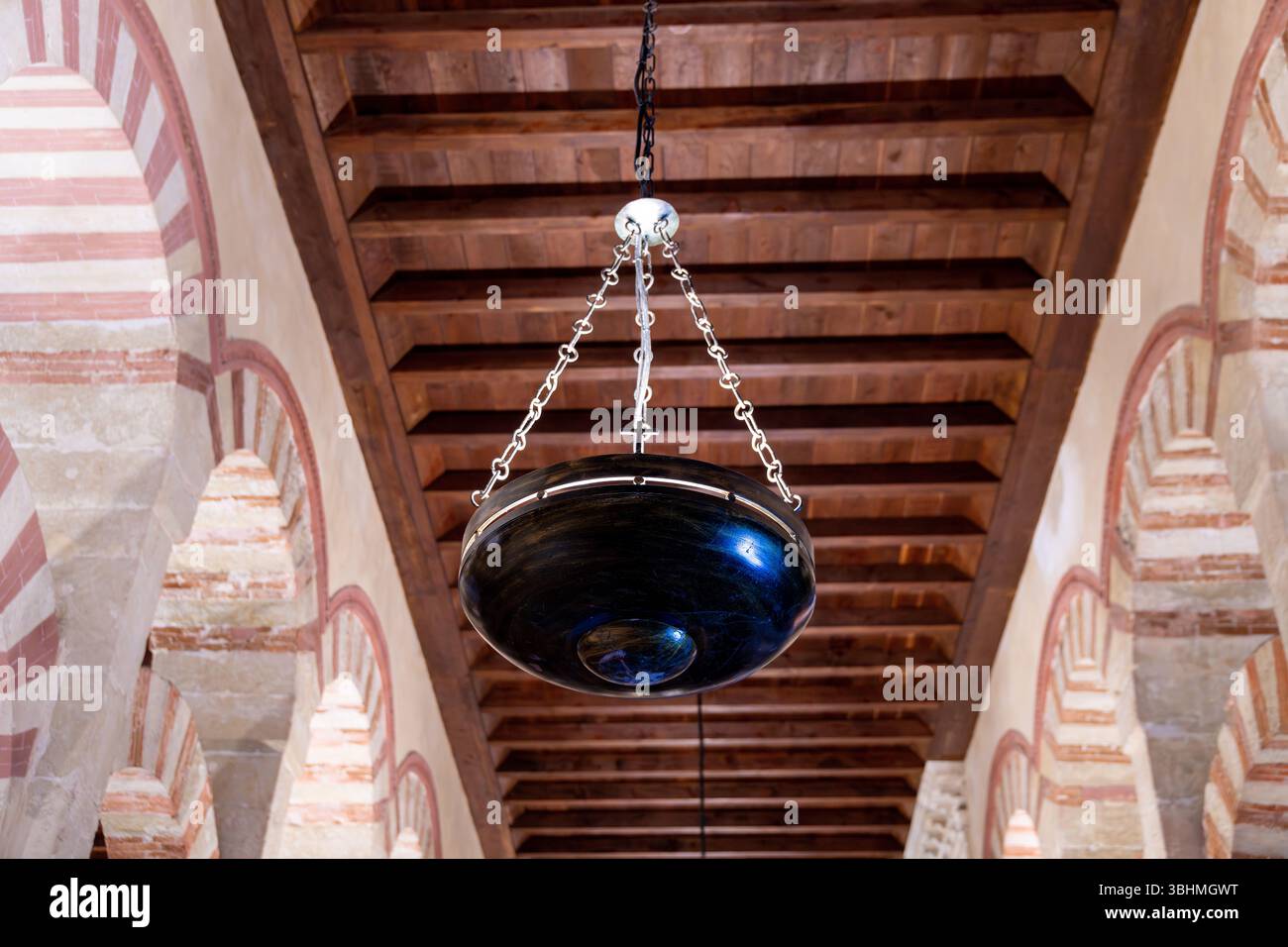 Cordova, Spagna - 08-04-2024: Una grande lampada in metallo scuro appesa a una catena su un soffitto di legno, circondata da archi a ferro di cavallo rossi e bianchi in una moschea- Foto Stock