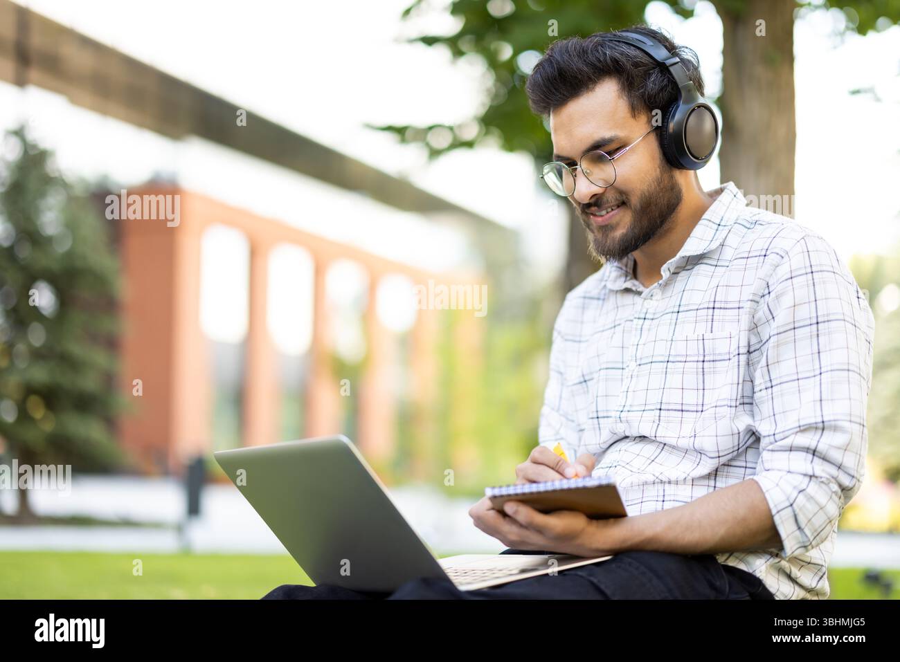 Un giovane uomo che lavora su un notebook all'aperto, indossa le cuffie e scrive su un notebook. Lo sfondo mostra alberi e un edificio. Foto Stock