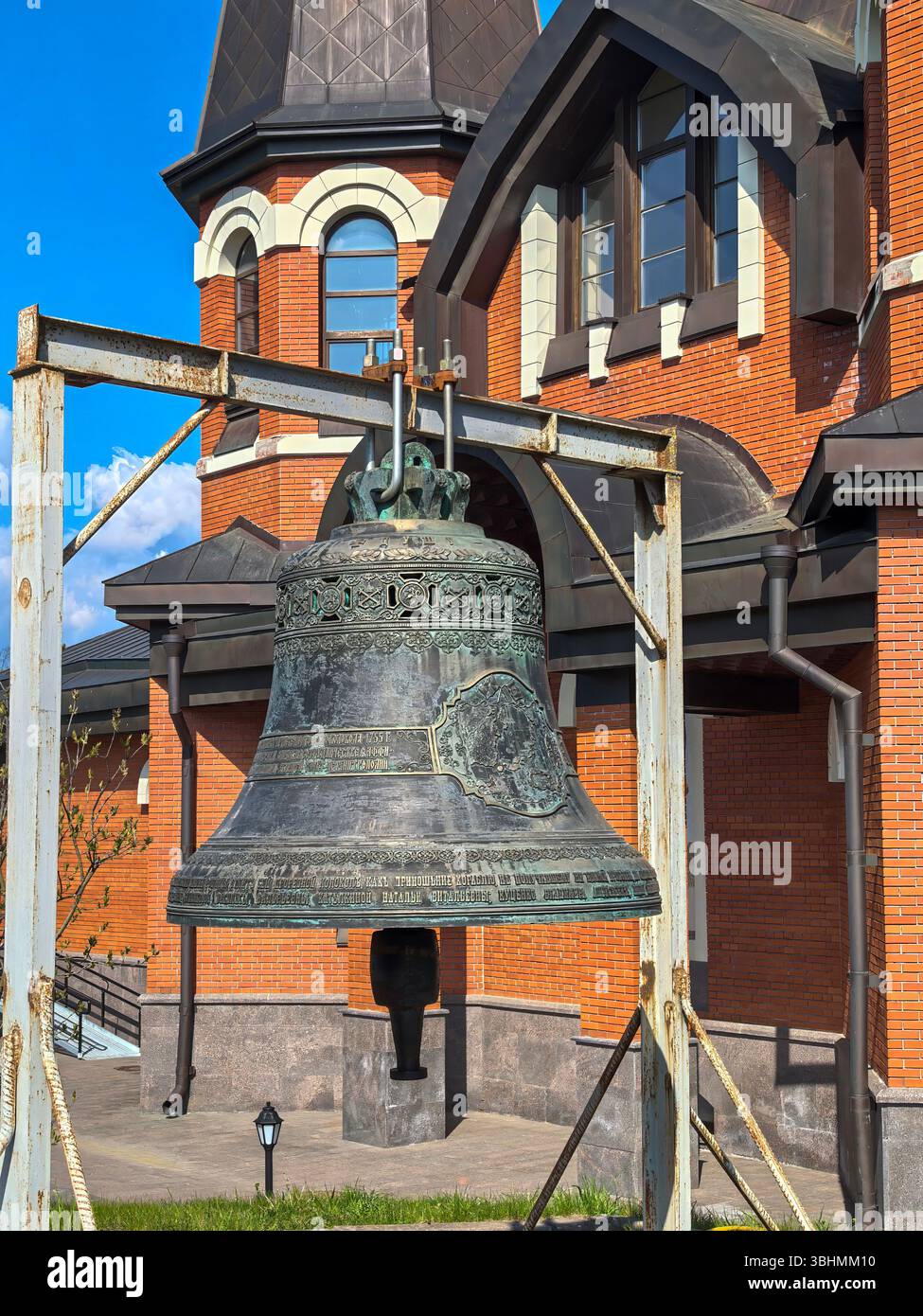 Yukki, Oblast' di Leningrado, Russia - 28 maggio 2025. Una grande campana dettagliata in primo piano, con la chiesa di San Giovanni Battista a Yukki Foto Stock