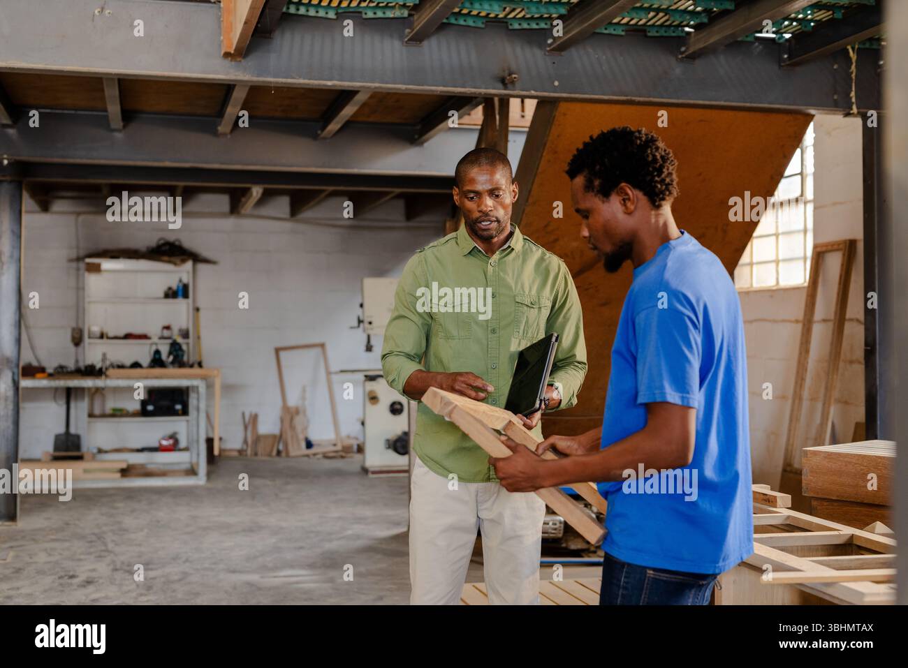 Colleghi afroamericani che esaminano la tavola di legno mentre consultano il tablet sul banco di lavoro dell'officina Foto Stock