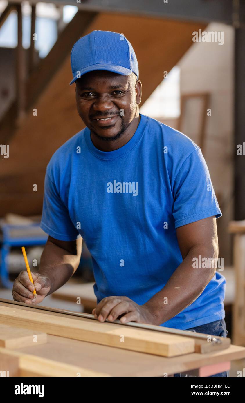 Uomo afro-americano che indossa T-shirt blu e tavola di marcatura del berretto con righello e matita in officina Foto Stock