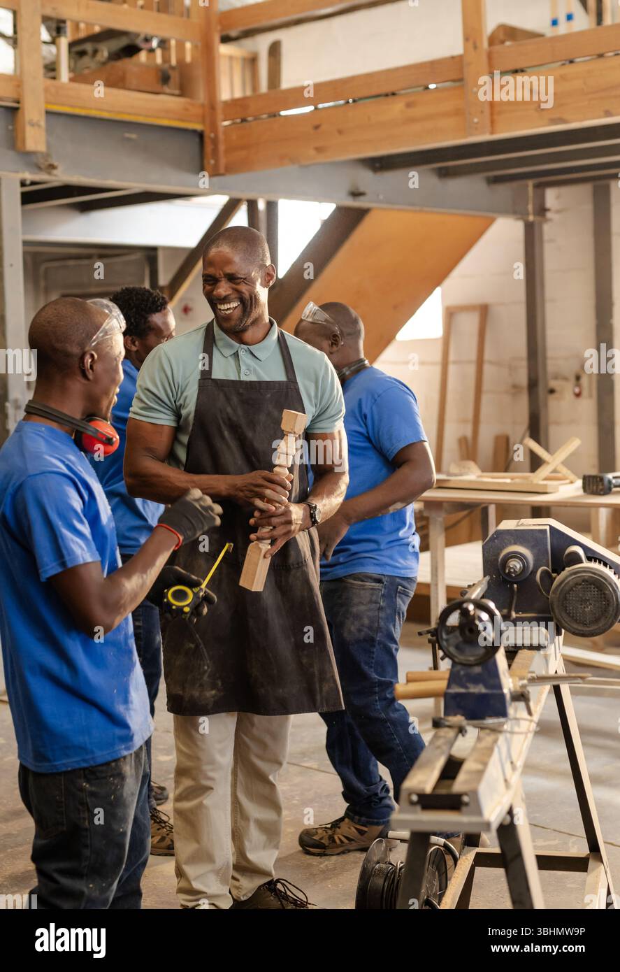 Squadra di falegnameria afroamericana che ride tenendo il mandrino intagliato accanto al tornio in legno in officina Foto Stock